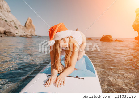 Woman sea sup. Close up portrait of happy young caucasian woman with long hair in Santa hat looking at camera and smiling. Cute woman portrait in a white bikini posing on sup board in the sea Woman sea sup. Close up portrait of happy young caucasian woman with long hair in Santa hat looking at camera and smiling. Cute woman portrait in a white bikini posing on sup board in the sea 111039189