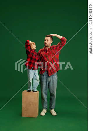 Child on pedestal and father standing beside, both posing with hands on heads against green studio background. Concept of International Day of Happiness 111040359