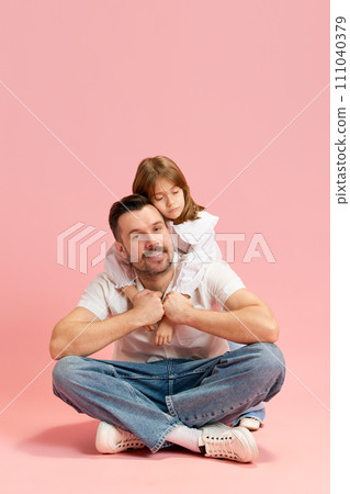 Content man sitting cross-legged with child resting on his back against pink pastel background. Concept of International Day of Happiness 111040379