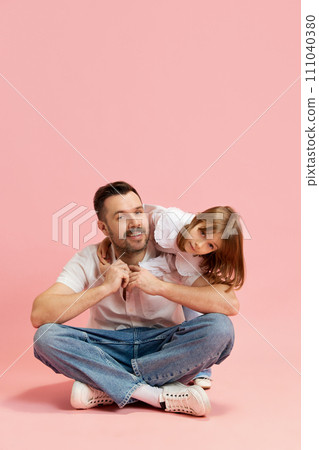 Young man sitting and little girl playing with him, Warm and lovely relationship between dad and daughters against pink background. Concept of International Day of Happiness Young man sitting and little girl playing with him, Warm and lovely relationship between dad and daughters against pink background. Concept of International Day of Happiness 111040380