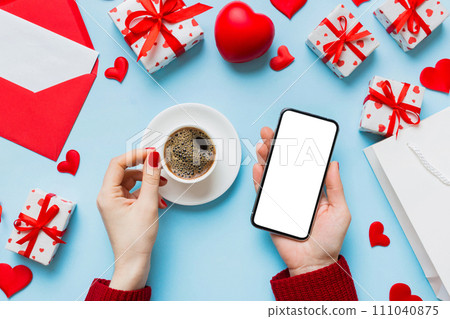 Female hands holding smart phone with coffee for Valentine day, gift box and envelope, hearts 111040875