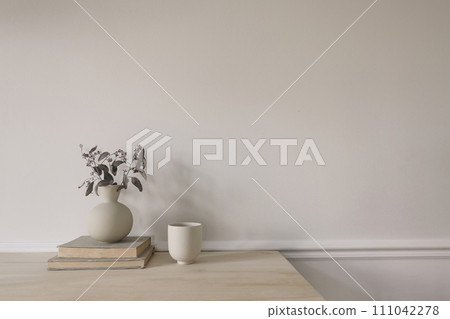 Breakfast, scandi interior still life. Minimal home design concept. Beige ceramic vase with dry eucalyptus tree branches. Cup of coffee on wooden table, desk with old books. Empty beige wall mockup. 111042278