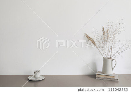 Autumn breakfast still life scene. Ceramic jug with dry grass and old books. Cup of coffee on beige table. Blank white wall mockup. Scandinavian interior, boho home. No people. Neutral design Autumn breakfast still life scene. Ceramic jug with dry grass and old books. Cup of coffee on beige table. Blank white wall mockup. Scandinavian interior, boho home. No people. Neutral design 111042283