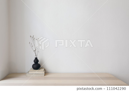 Elegant interior still life, minimal home design concept. Organic shaped black vase with dry grass on table, desk with old books. Working space, home office decor. Empty beige wall mockup. 111042290