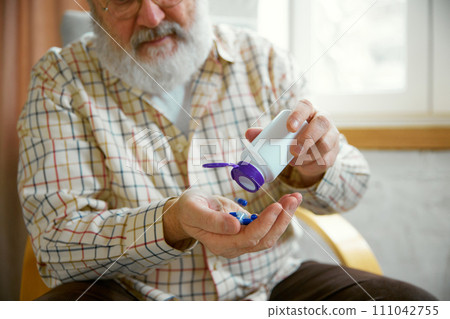 Senior man with beard pouring pills into his hand from a container, seated indoors. Drinking vitamins and feeling good Senior man with beard pouring pills into his hand from a container, seated indoors. Drinking vitamins and feeling good 111042755
