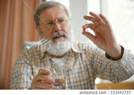 Senior man siting at home and drinking pills. Blurred background. Focus on hands with red capsule, pill. Vitamins Senior man siting at home and drinking pills. Blurred background. Focus on hands with red capsule, pill. Vitamins 111042757