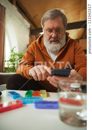 Elderly man with glasses using smartphone, checking medical prescriptions, contacting with doctor. Pill organizer and water glass on table 111042817