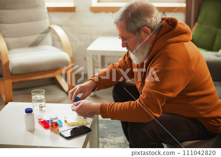 Elderly person taking pill into hand, with water glass, smartphone, and pill organizer in the background. Daily vitamins and meds Elderly person taking pill into hand, with water glass, smartphone, and pill organizer in the background. Daily vitamins and meds 111042820