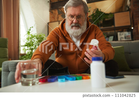 Senior man taking care after health with meds, vitamins. Man sitting at home, taking glass of water for pills 111042825