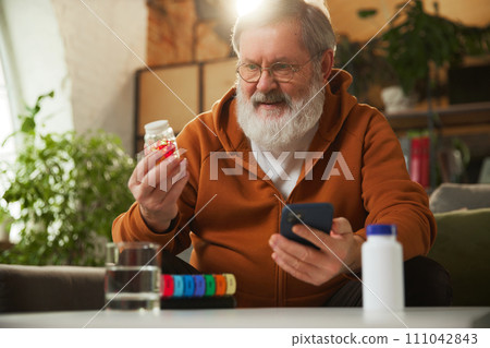 Senior bearded man examining pill bottle while looking at smartphone. Using pill organizer. Online med services, checking prescription Senior bearded man examining pill bottle while looking at smartphone. Using pill organizer. Online med services, checking prescription 111042843