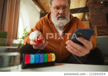 Elderly man examining pill bottle while looking at smartphone. Using pill organizer. Online med services Elderly man examining pill bottle while looking at smartphone. Using pill organizer. Online med services 111042854