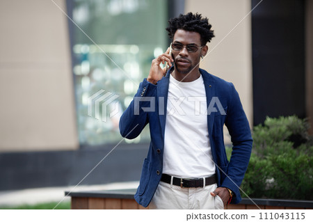Confident African American businessman in sharp suit and casual tee, speaking on mobile phone and walking in urban area. Trendy portrayal of modern city life Confident African American businessman in sharp suit and casual tee, speaking on mobile phone and walking in urban area. Trendy portrayal of modern city life 111043115