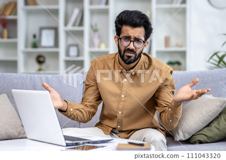 Portrait of upset Indian young man who works from home, sitting on couch at table with laptop and documents, looking at camera and throwing up hands in frustration. 111043320