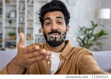 Close-up photo of a smiling young Indian man at home and talking on a video call on the phone to the camera, gesturing with his hands. Close-up photo of a smiling young Indian man at home and talking on a video call on the phone to the camera, gesturing with his hands. 111043321