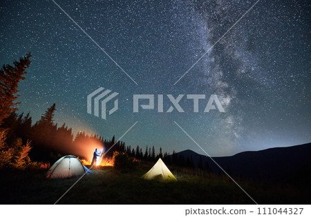 Male photographer taking pictures of Milky way. Man doing photo shoot of starry sky next to his camping. Starry night in mountains. 111044327