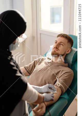 Nurse in hijab removing catheter from vein in arm of male blood donor after procedure, focus on man Nurse in hijab removing catheter from vein in arm of male blood donor after procedure, focus on man 111044725