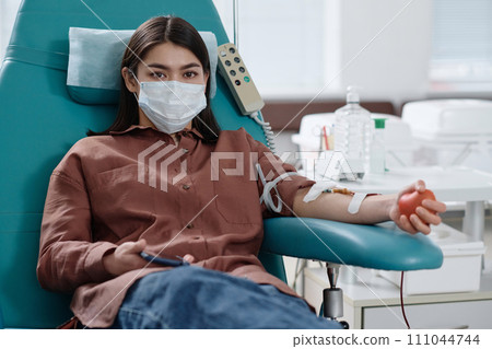 Young woman in protective mask looking at camera during process of blood donation in medical center Young woman in protective mask looking at camera during process of blood donation in medical center 111044744