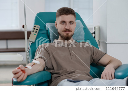 Young caucasian man sitting in armchair in clinic during blood transfusion and smiling at camera Young caucasian man sitting in armchair in clinic during blood transfusion and smiling at camera 111044777