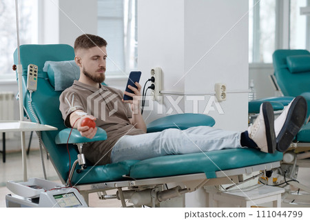Male blood donor sitting in armchair during transfusion process in clinic and browsing smartphone Male blood donor sitting in armchair during transfusion process in clinic and browsing smartphone 111044799