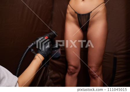 Cosmetologist using airbrush for spray tan apply to young woman body in beauty salon. Cosmetologist using airbrush for spray tan apply to young woman body in beauty salon. 111045015