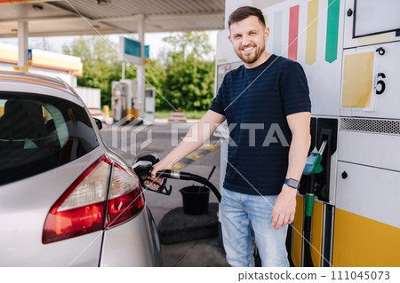 Bearded man refuelling car on gas station and looking into his smartphone. Man compares fuel prices Bearded man refuelling car on gas station and looking into his smartphone. Man compares fuel prices 111045073