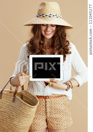 smiling female in blouse and shorts showing tablet PC on beige 111045277