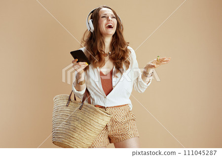 smiling stylish woman in blouse and shorts using phone on beige 111045287