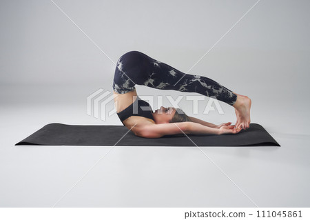 Motivated Caucasian female resting in Halasan pose on yoga mat. Side view of slender, fit woman in black pants and top performing plow pose on floor, isolated on grey. Concept of yoga, lifestyle. Motivated Caucasian female resting in Halasan pose on yoga mat. Side view of slender, fit woman in black pants and top performing plow pose on floor, isolated on grey. Concept of yoga, lifestyle. 111045861