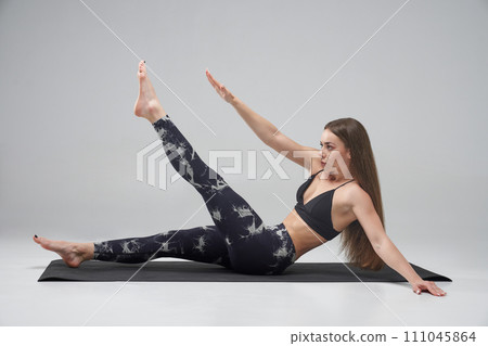 Sportswoman with long fair hair doing crunches in studio. Side view of young Caucasian female raising arm and leg, while having abs workout, isolated on grey background. Concept of sporty lifestyle.  111045864