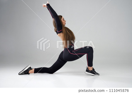 Flexible female in activewear stretching body in Crescent Lunge pose of yoga in studio. Side view of Caucasian woman with long hair practicing yoga asana on floor, isolated on grey. Yoga concept. Flexible female in activewear stretching body in Crescent Lunge pose of yoga in studio. Side view of Caucasian woman with long hair practicing yoga asana on floor, isolated on grey. Yoga concept. 111045876