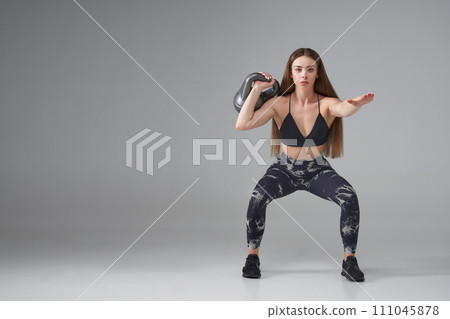 Determined Caucasian woman in legging, doing squats with kettle bell indoors. Front view of fitness girl using kettle bell, while exercising, isolated on grey background. Sport, body building concept. Determined Caucasian woman in legging, doing squats with kettle bell indoors. Front view of fitness girl using kettle bell, while exercising, isolated on grey background. Sport, body building concept. 111045878