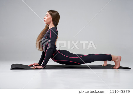 Focused young woman in activewear stretching body indoors. Side view of flexible female yoga trainer with fair hair training on yoga mat, isolated on grey studio background. Concept of yoga, pilates. 111045879
