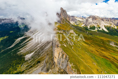 Dense foggy cloud descends on giant Seceda ridgeline with green slopes and bare peak. Cloudy sky above touristic Italian Alps aerial view Dense foggy cloud descends on giant Seceda ridgeline with green slopes and bare peak. Cloudy sky above touristic Italian Alps aerial view 111047271