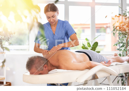 Skilled middle-aged masseuse conducting back massage for man client Skilled middle-aged masseuse conducting back massage for man client 111052184