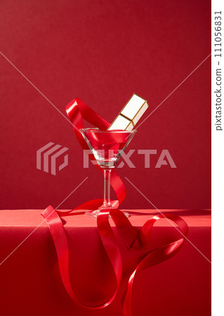 A lipstick and red silk ribbon inside a cocktail glass on a red background. Romantic gift for Valentine Day. The space is viewed from the front for advertising. 111056831
