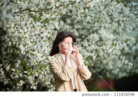 Woman allergic suffering from seasonal allergy at spring in blossoming garden at springtime. Young woman sneezing and blowing nose with nasal handkerchief in front of blooming tree. Allergy concept. 111058596