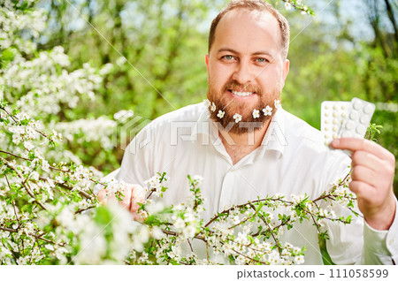 Man allergic suffering from seasonal allergy at spring. Handsome man with small white flowers dotted in beard, holding pack of pills, posing in blossoming garden. Antihistamine medication concept 111058599