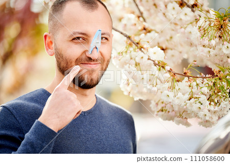 Man allergic suffering from seasonal allergy at spring. Bearded man with clothespin clipped to his nose - symbolic gesture of his inability to breathe due to nasal congestion near blooming tree. 111058600