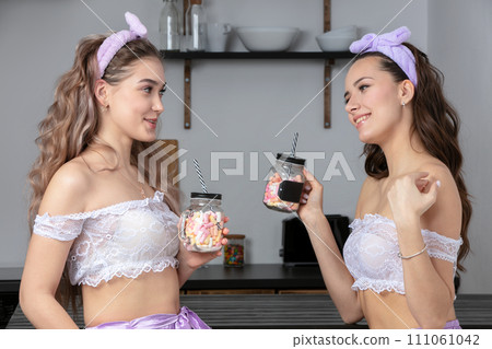 Two beautiful girls in the kitchen. Girlfriends in pajamas with sweets at home. Young women are talking. Two beautiful girls in the kitchen. Girlfriends in pajamas with sweets at home. Young women are talking. 111061042