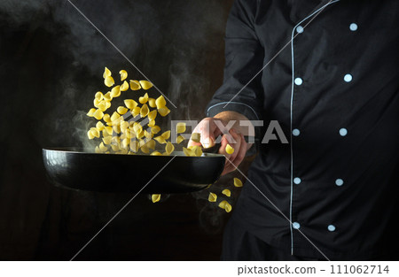 The cook throws the pasta into a hot, steamy frying pan. Italian cuisine or shell-type pasta. Place for recipe on black background The cook throws the pasta into a hot, steamy frying pan. Italian cuisine or shell-type pasta. Place for recipe on black background 111062714