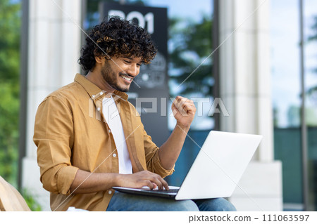 A young Muslim man sits on a bench with a laptop on the city street, looks at the screen and rejoices in success, showing a victory gesture with his hand. A young Muslim man sits on a bench with a laptop on the city street, looks at the screen and rejoices in success, showing a victory gesture with his hand. 111063597