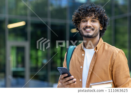 Close-up portrait of young smiling Muslim male student holding books and mobile phone and looking at camera. 111063625