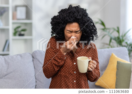 Young woman feeling unwell, sneezing into tissue, holding mug at home. Concepts of sickness, flu, cold, allergy, and healthcare. 111063680