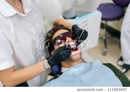 Closeup of unrecognizable female dentist using modern diode dental laser for periodontal care. Male patient wearing protective glasses, preventing eyesight damage. Periodontitis, dental hygiene. 111063697