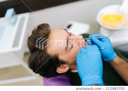 Closeup face of male patient sitting in stomatology seat with open mouth during unrecognizable male dentist examining teeth, with dental tool in dentistry clinic. Concept of stomatology. Closeup face of male patient sitting in stomatology seat with open mouth during unrecognizable male dentist examining teeth, with dental tool in dentistry clinic. Concept of stomatology. 111063702