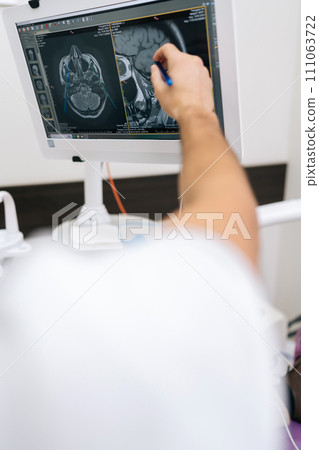 Vertical shot of male dentist pointing at teeth radiography on monitor to explain dentition diagnosis to young man patient with toothache. Dental expert showing x-ray scan results to man. Vertical shot of male dentist pointing at teeth radiography on monitor to explain dentition diagnosis to young man patient with toothache. Dental expert showing x-ray scan results to man. 111063722