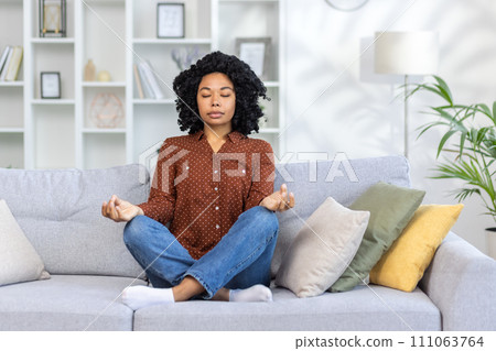 Concentrated and calm African American woman sitting on sofa at home in lotus position, doing yoga and meditating with eyes closed. Concentrated and calm African American woman sitting on sofa at home in lotus position, doing yoga and meditating with eyes closed. 111063764