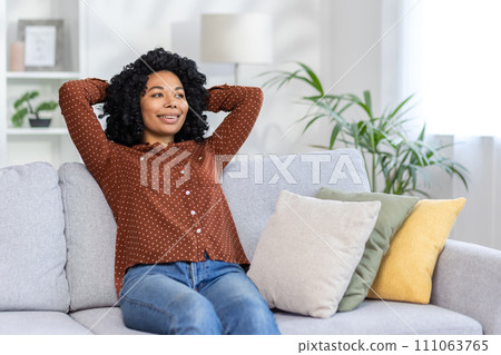 Smiling young African American woman sitting on sofa at home and relaxing with self satisfaction. threw her hands behind her head and looks to the side. Smiling young African American woman sitting on sofa at home and relaxing with self satisfaction. threw her hands behind her head and looks to the side. 111063765