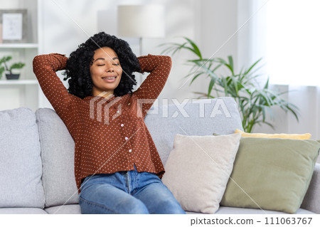 Relaxed African American woman enjoying a peaceful moment, reclining on a couch at home, eyes closed with a content smile. 111063767