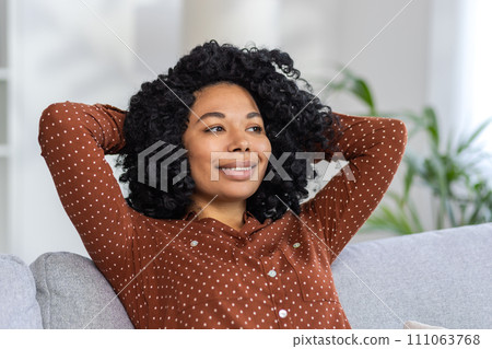 Close-up photo of a smiling African-American woman sitting on the couch at home with her hands behind her head, and confidently nodding to the side. Close-up photo of a smiling African-American woman sitting on the couch at home with her hands behind her head, and confidently nodding to the side. 111063768
